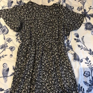 Blommig klänning med korta ärmar - Charmig blommig klänning i mörkblått/svart med vita blommor. Den har korta ärmar och en luftig passform, perfekt för en avslappnad stil. Klänningen är tillverkad i ett lätt och bekvämt material.