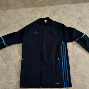 Mörkblå vindjacka från Adidas - Säljer en mörkblå vindjacka från Adidas med ljusblå ränder längs ärmarna och sidorna. Jackan har dragkedja framtill och två fickor med dragkedjor. Perfekt för sportiga aktiviteter eller en avslappnad stil.