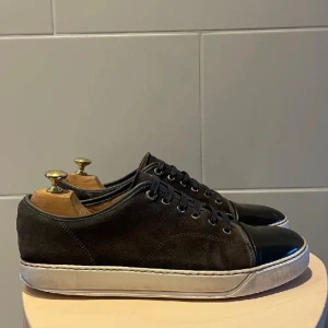 Lanvin uk9 - Snygga svarta sneakers med en stilren design och lackdetaljer på tån. Skorna har en vit sula och snörning framtill. Perfekta för en casual look med en touch av elegans. Lite slitningar vid insidan av hälen, men det är inga besvär man känner av när man väl bär skorna uk9
