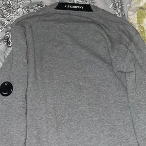 Grå sweatshirt från C.P. Company - Säljer en stilren grå sweatshirt från C.P. Company. Tröjan har en rund halsringning och en unik detalj på ärmen. Perfekt för en avslappnad look.