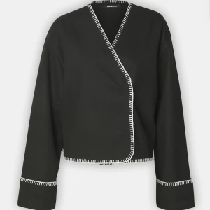 Svart omlottjacka med vit kant - Snygg svart omlottjacka från Gina Tricot med vit kantdetalj. Jackan har en stilren design med långa ärmar och en unik omlottstil. Perfekt för en elegant look.