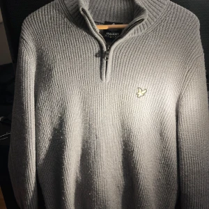 Grå tröja med dragkedja från Lyle & Scott - Säljer en stilren grå tröja från Lyle & Scott med dragkedja och broderad logga på bröstet. Tröjan är ribbad och har en hög krage, perfekt för kyligare dagar. Passar både till jeans och chinos för en avslappnad look.