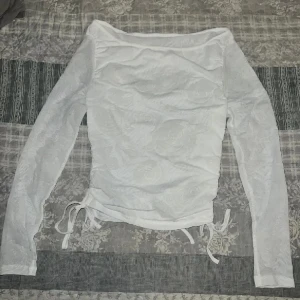 Vit långärmad topp med blommönster - Säljer en vit långärmad topp med subtilt blommönster. Toppen har knytband i nederkant för justerbar passform. Perfekt för en stilren look.
