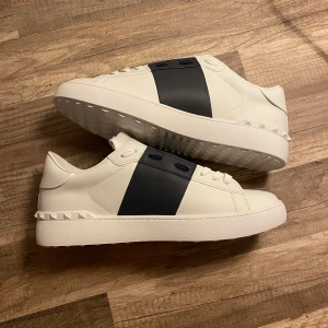 Valentino sneakers med svart detalj - Snygga vita sneakers från Valentino med en bred svart rem över ovansidan. Skorna har en stilren design med en platt sula och subtila nitar runt hälen. Perfekta för en grischig look.