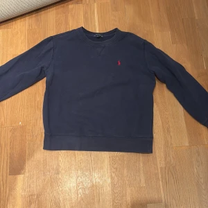 Mörkblå collage tröja från Ralph Lauren - Säljer en stilren mörkblå tröja från Ralph Lauren med det klassiska röda logotypbroderiet på bröstet. Tröjan har långa ärmar och en rund halsringning. Perfekt för en avslappnad och snygg look.