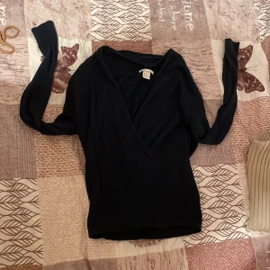 Svart omlottopp från H&M - Snygg svart omlottopp från H&M med långa ärmar. Perfekt för en stilren look. Passar bra till både jeans och kjol. Ett måste i garderoben för den som gillar enkel elegans.