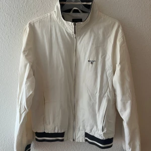 Vit jacka från GANT - Säljer en vit jacka från GANT med dragkedja och broderad logga på bröstet. Jackan har ribbade muddar i svart och vit randigt mönster. Perfekt för en sportig look 