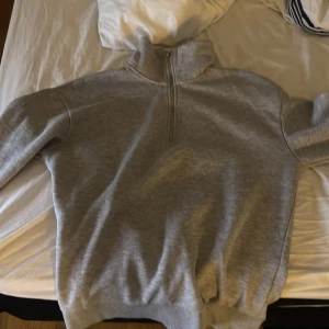 Grå tröja med dragkedja från RYVLS - Säljer en stilren grå tröja från RYVLS med halv dragkedja och hög krage. Perfekt för kyligare dagar med sin mjuka insida. Tröjan har långa ärmar och ribbade muddar vid ärmslut och nederkant. Passar bra till både jeans och träningsbyxor.