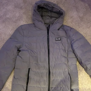 Grå dunjacka från Emporio Armani - Snygg grå dunjacka från Emporio Armani med huva och dragkedja framtill. Jackan har en quiltad design och är perfekt för kyligare dagar. Den har också praktiska fickor med dragkedja.