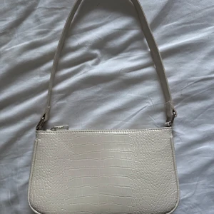 Vit axelväska - Snygg vit axelväska med krokodilmönster och dragkedja. Perfekt för att ge en stilren touch till din outfit på fest eller i vardagen. Väskan har ett rymligt fack med svart foder med två praktiska innerfickor och ett fack med dragkedja i. Enbart använd 1 gång.