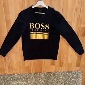 Svart tröja från Hugo Boss med gulddetaljer - Säljer en svart tröja från Hugo Boss med guldfärgad logga på bröstet. Tröjan har långa ärmar och är perfekt för en stilren look. Passar bra till både jeans och chinos. Har hål där back men det syns ej köpt för 1800kr på room 1006 malmö