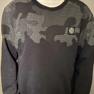 Svart tröja med kamouflagemönster från Stone Island - Säljer en svart tröja från Stone Island med ett coolt kamouflagemönster i grått. Tröjan har en rund halsringning och långärmade ärmar. Perfekt för dig som gillar stilrena och unika plagg. Den har krumt lite i tvätten så den är s-m 