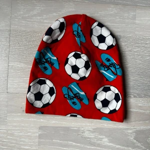 Orange mössa med fotbollsmönster - Säljer en cool orange mössa med tryck av fotbollar och blå fotbollsskor. Perfekt för fotbollsälskare och att hålla sig varm under kyliga dagar. Strl 2