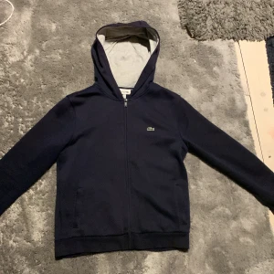 Mörkblå hoodie från Lacoste Sport - Säljer en stilren mörkblå hoodie från Lacoste Sport med dragkedja och huva. Skick 8/10