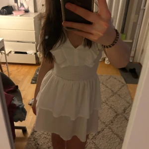 Vit klänning med volangdetaljer - Charmig vit klänning med volangdetaljer och v-ringning. Klänningen har korta ärmar och en smockad midja som ger en fin passform. Perfekt för somriga dagar.