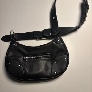 Svart axelväska från H&M -  Svart axelväska i läderimitation från h&m. Väskan har en justerbar rem med ett stort spänne och en praktisk dragkedja framtill. Endast använd 2 gånger🤗
