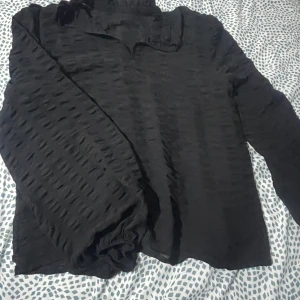 Svart blus från NA-KD - Säljer en elegant svart blus från NA-KD med långärmad design. Blusen har en unik textur och elastiska ärmslut för en bekväm passform. Perfekt för att ge en stilren touch till din outfit.