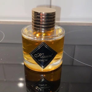 Kismet Magic Parfym - Endast testsprutad. Elegant parfymflaska med en gyllene vätska och en stilren, rund design. Flaskan har en glänsande guldfärgad kork och en svart etikett med texten 'Kismet Magic'. Perfekt för den som söker en magisk doftupplevelse.