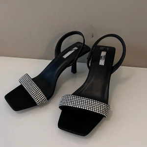 Svarta klackar med strassdetaljer från Primark - Snygga svarta klackar från Primark i storlek 39. De har en elegant klack och en rem med glittriga strassdetaljer som ger en lyxig känsla. Perfekta för festliga tillfällen eller en utekväll. Skorna är i nytt skick och redo att bäras med stil!