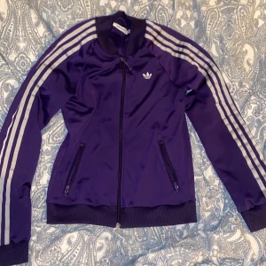 Lila träningsjacka från Adidas - Säljer en otroligt snygg lila träningsjacka från Adidas i mycket bra skick. Jackan har de klassiska tre ränderna längs ärmarna och en dragkedja framtill. Perfekt för träning eller som en cool vardagslook. Storlek 38, passar perfekt för dig som gillar en normal passform. 