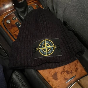 Stone Island mössa - Säljer en svart ribbad mössa från Stone Island. Den är i mycket bra skick och har en broderad logga framtill. Mössan är gjord av bomull och ull, perfekt för höst och vinter. Den är både stilren och varm, idealisk för kalla dagar. Aldrig använd