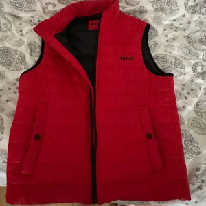 Röd dunväst från Hugo - Snygg röd dunväst från Hugo i mycket bra skick. Västen har en dragkedja framtill och två fickor med knappar. Perfekt för höst och vår, och den är quiltad för extra värme. Passar bra för både vardag och festliga tillfällen.