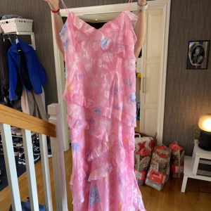 Rosa blommig långklänning - Säljer en superfin rosa långklänning med blommigt mönster. Klänningen har smala axelband och volangdetaljer som ger en romantisk känsla. Perfekt för sommarens fester eller en mysig midsommar. Materialet känns lätt och luftigt, vilket gör den idealisk för varma dagar. 🌸 storlek 46 men mer som en 42/44 skulle jag säga🥰