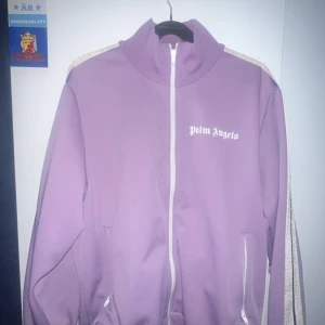 Lila jacka från Palm Angels - Säljer en snygg lila jacka från Palm Angels i storlek L. Jackan har en dragkedja framtill och vita detaljer på ärmarna. Perfekt för en cool och avslappnad stil. Passar bra till både vardag och fest! Har kvitto 