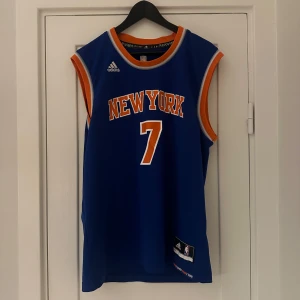 Blå New York basketlinne från Adidas - Säljer ett blått basketlinne från Adidas med New York-tryck och nummer 7 på framsidan. På baksidan står det Anthony och samma nummer. Linnet har orange och vita detaljer vid ärmhålen och halsen. Perfekt för sport eller som en cool vardagslook! 🏀