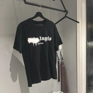 Svart t-shirt från Palm Angels - Säljer en svart t-shirt från Palm Angels i mycket bra skick. Den har ett coolt tryck med texten 'Palm Angels' i vitt på framsidan. Perfekt för en avslappnad stil eller att bära på fest. Passar bra till jeans eller shorts!