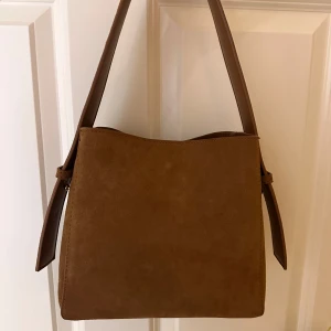 Brun axelväska i mocka - Snygg brun axelväska i mjuk mocka. Perfekt för vardagsbruk med justerbar axelrem och stilren design. Väskan har en enkel men elegant look som passar till alla outfits.