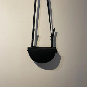 Svart axelväska i skinn - Snygg och stilren svart axelväska i skinn med justerbar rem. Perfekt för både vardag och fest! Väskan har en halvmåneform och är enkel att matcha med olika outfits. Ett måste i garderoben för den som gillar minimalistisk stil.