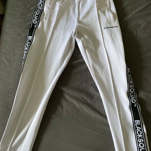 Vita byxor från BlackSquad - Säljer ett par vita byxor från BlackSquad med svarta sidoremsor där märket står tryckt. Byxorna har en normal passform och är perfekta för en avslappnad stil. De har dragkedja och snörning i midjan för bästa komfort. Perfekta för både vardag och träning!