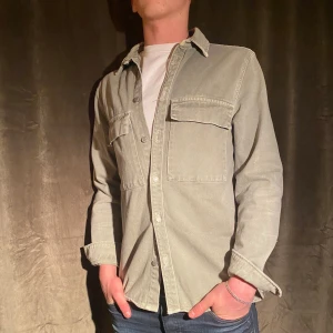 Grön Overshirt  - Säljer en snygg grön overshirt från Zara i storlek M. Den har en cool vintage-look med två stora bröstfickor och knappar framtill. Perfekt för lager-på-lager-stil under hösten eller våren. Materialet känns robust och är perfekt för en avslappnad men stilren look. Passar både till vardags och lite mer uppklädda tillfällen. Vid minsta fråga eller fundering hör av er🤝