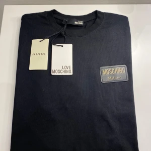 Svart t-shirt från Love Moschino - Säljer en snygg svart t-shirt från Love Moschino. Den har en stilren design med en patch på bröstet där det står 'Moschino Milano'. Perfekt för både vardag och fest! Storlek XL men kan passa L 