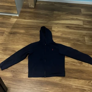 Blå hoodie från Polo Ralph Lauren - Säljer en mörkblå hoodie från Polo Ralph Lauren i storlek 14-16 . Den har en dragkedja framtill och är perfekt för både höst och vinter. Hoodien är i bra skick och har en klassisk design med en liten logga på bröstet. Nypris: 1200kr