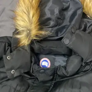 Säljer en svart dunjacka från Canada Goose i storlek XXL. Jackan har en avtagbar huva med snygg pälsdetalj och dragkedja framtill. Perfekt för kalla vinterdagar! 🖤