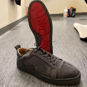 Gråa loubutins utan spikar  - Snygga grå sneakers i mocka med den ikoniska röda sulan. Perfekta för både vardag och fest. Skorna har snörning och en stilren design som passar till det mesta. De är i mycket bra skick och redo att användas direkt. Ett måste för den stilmedvetna!