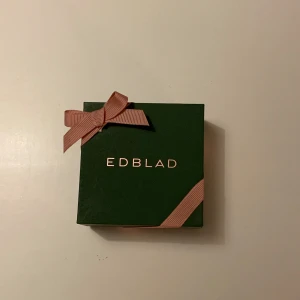 Halsband med hjärthänge från Edblad - Säljer ett elegant halsband från Edblad med ett hjärthänge. Hänget är i guld och dekorerat med glittrande stenar, perfekt för att ge en touch av glamour till vilken outfit som helst. Kommer i en fin grön presentask med rosa rosett. Perfekt som present eller till dig själv!