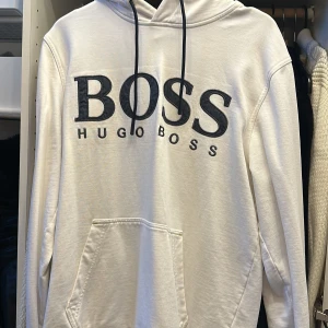 Vit hoodie från Hugo Boss - Säljer en stilren vit hoodie från Hugo Boss i storlek M. Den har en stor BOSS-logga på framsidan och en praktisk känguruficka. Perfekt för en avslappnad stil och passar till alla säsonger. Huvan har justerbara snören för bästa passform. 