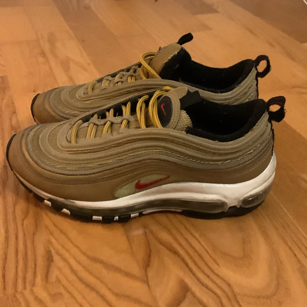 Säljer ett par Nike Air Max 97 i en snygg guld och svart färgkombination. Skorna har den klassiska vågiga designen med en röd Nike-logga på sidan. De är i bra skick med synlig Air-dämpning i sulan för extra komfort. Perfekta för både vardag och fest!. Skor.