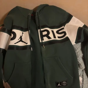 Grön hoodie från Jordan - Säljer en snygg grön hoodie från Jordan med dragkedja och coolt tryck med texten 'Paris' och den ikoniska Jumpman-loggan. Perfekt för vardagsbruk eller sportiga tillfällen. Den har långa ärmar och en bekväm passform. Passar perfekt till höst och vår! Köpt för nästan 1500