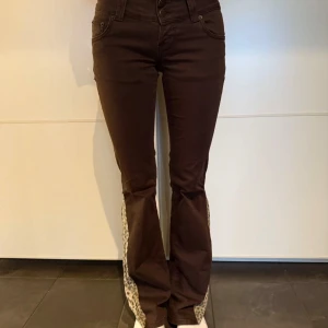 Bruna bootcut byxor med leoparddetaljer - Säljer ett par snygga bruna bootcut byxor med leopardmönstrade detaljer längs sidorna. Byxorna har låg midja och är dekorerade med pärlor på bakfickorna. Perfekta för en unik och trendig look! Midja: 34,5 cm Innerbenslängd: 82 cm Ytterbenslängd: 105 cm