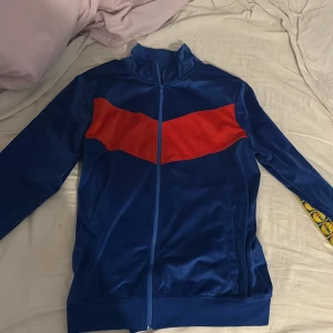 Blå och röd träningsjacka från Lidl  - Säljer en blå träningsjacka med röd detalj på bröstet och gula mönster längs ärmarna. Jackan har dragkedja och är långärmad, perfekt för träning eller vardagsbruk. Den är i bra skick och har en skön passform. Skriv vid minsta fråga!