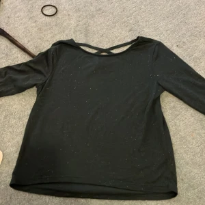 Mörkgrön långärmad topp från H&M - Säljer en svart långärmad topp från H&M i storlek 146/152. Den har en snygg detalj med korsade band i ryggen och är perfekt för både vardag och fest. Materialet är mjukt och bekvämt, med ett diskret glittrigt mönster som ger lite extra stil. Passar perfekt till jeans eller kjol!