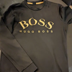 Hugo Boss sweatshirt - Säljer en stilren svart tröja från Hugo Boss med guldtext på bröstet. Tröjan har långa ärmar och en bekväm passform. Perfekt för både vardag och fest. Passa på att köpa denna snygga tröja!