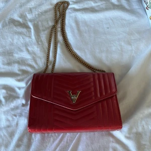 Röd quiltad axelväska - Snygg röd axelväska med quiltat mönster och guldfärgad kedja. Väskan har en elegant V-detalj på framsidan och är perfekt för både vardag och fest. Den är i mycket bra skick och ger en lyxig touch till vilken outfit som helst.