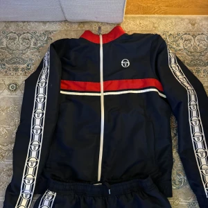 Svart träningsdräkt från Sergio Tacchini - Säljer en svart träningsdräkt från Sergio Tacchini i mycket bra skick. Dräkten har vita och röda detaljer med märkets logga längs ärmar och ben. Perfekt för sportiga aktiviteter eller en avslappnad stil. Dragkedja framtill och elastisk midja för bekväm passform. Dressen är ganska tjock så den är varm perfekt för vintern! Ps.stor i storlek 
