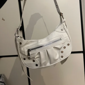 Vit axelväska med dragkedja och nitar - Säljer en snygg vit axelväska i syntetmaterial. Väskan har en cool design med dragkedja framtill och dekorativa nitar. Perfekt för att ge din outfit en edgy touch. Justerbar axelrem för bästa passform. Använd 2 ggr.