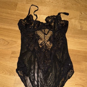 Svart spetsbody - Säljer en elegant svart spetsbody med vackra detaljer. Den är ärmlös och har en fin spetsdesign framtill som ger en lyxig känsla. Perfekt för en festlig kväll eller en speciell tillställning. Passar bra under en kavaj eller som den är. Justerbara axelband för bästa passform.